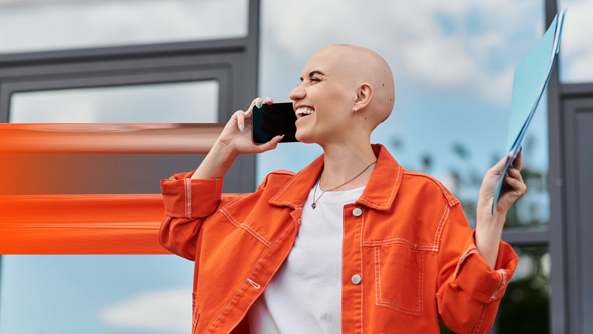 Young woman with alopecia smiling and talking on the phone outside a modern building By LightFieldStudios_L1