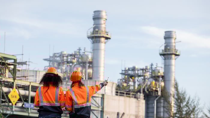 Two female engineers in orange safety jackets and helmets stand at an industrial plant site