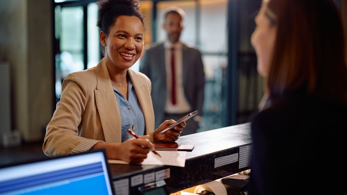 Happy black woman checking in at hotel reception during her business trip. By drazenphoto_L1